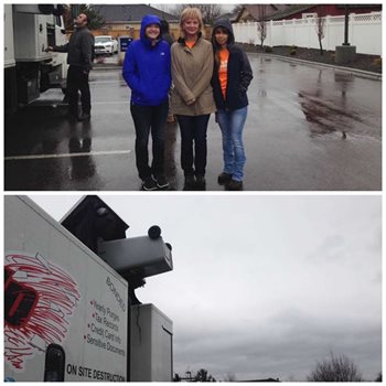 three women standing in a wet parking lot next to giant paper shredding machine