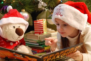 young girl wearing a santa hat reading a book to a stuff animal