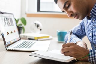 young man writing in a notebook while researching on a laptop