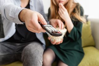 man and woman on couch, woman eating something and man clicking a button on a TV remote