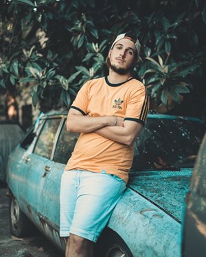 Young man with beard leaning against a beat up old blue car