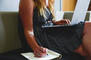 young woman with laptop on lap and writing in notebook next to her