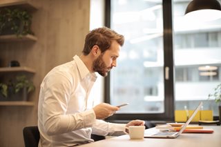 man in white shirt holding a smartphone and looking at a laptop
