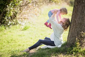 young mother holding her little girl over her head with a tree behind them