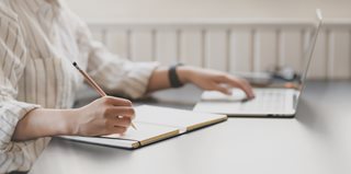 woman at a table with a laptop writing in a notebook