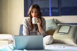 young woman sitting on a bed blowing on hot coffee and looking at her laptop