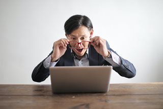 Asian man with glasses shocked looking at laptop