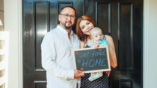 man and woman with a baby holding a sign that says "Our First Home"