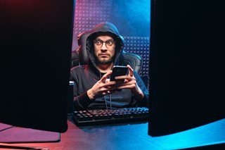 man in dark hoodie holding phone and looking at computer screens